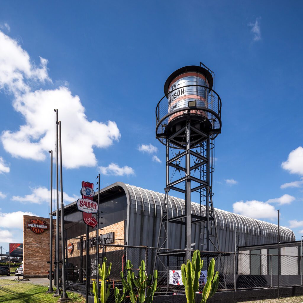 Uma concessionária Harley-Davidson com um prédio de tijolos e metal, uma grande torre de água com a inscrição "Harley-Davidson", placas de sinalização, um cacto em primeiro plano e um céu azul com nuvens dispersas ao fundo. - Luiz Volpato Arquitetura