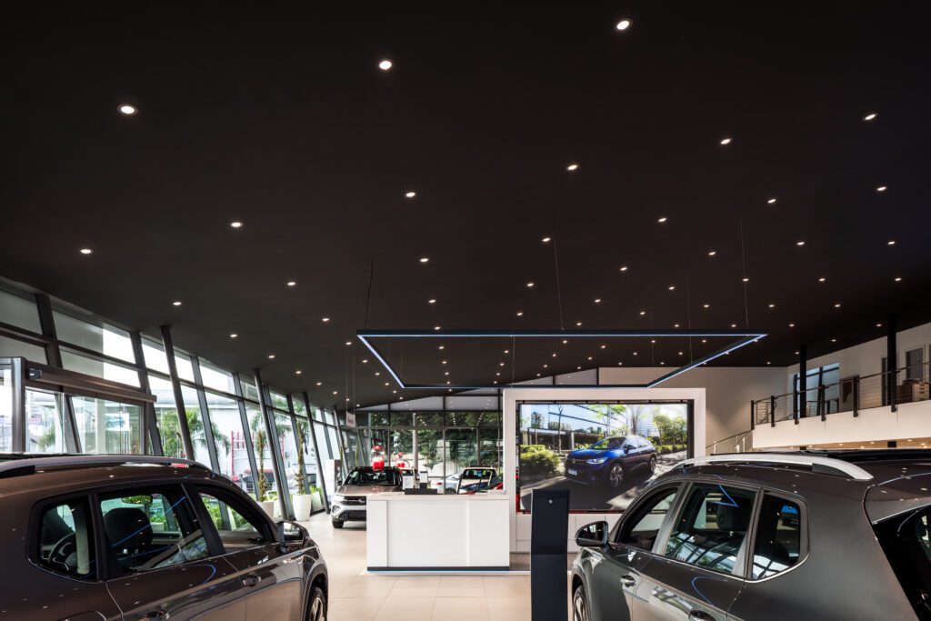 Um showroom moderno de uma concessionária de automóveis com janelas grandes, iluminação superior brilhante e dois carros em primeiro plano. Um display digital com a imagem de um carro está na parede do fundo, e há um balcão de recepção elegante e minimalista no centro. - Luiz Volpato Arquitetura