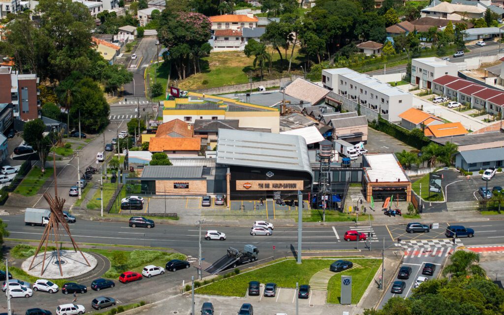 Vista aérea de uma rua urbana movimentada com vários carros estacionados, um monumento circular com postes de madeira e um grande edifício no centro exibindo o logotipo da Harley-Davidson. Ao redor, há casas, árvores e pequenas empresas sob um céu ensolarado. - Luiz Volpato Arquitetura