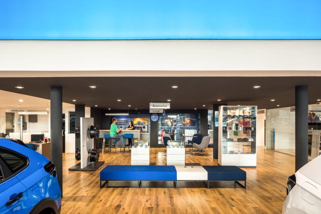 Um showroom de carros moderno com piso de madeira, iluminação clara e detalhes em azul. Os carros são parcialmente visíveis à esquerda e à direita. Vitrines com acessórios e mercadorias ficam no centro e uma pessoa trabalha em uma mesa nos fundos. - Luiz Volpato Arquitetura