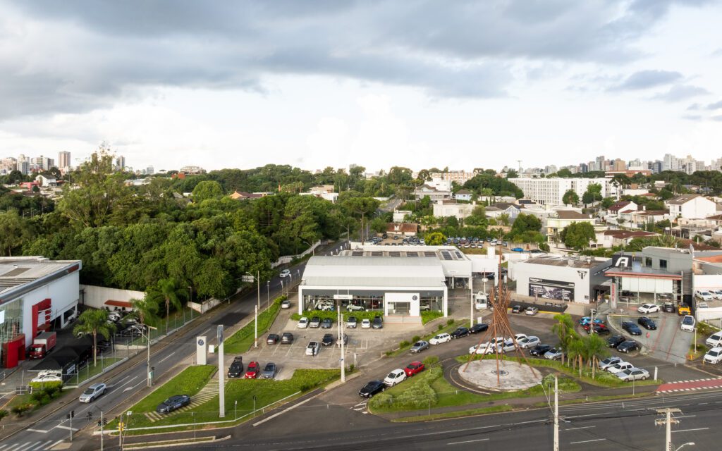 Vista aérea de uma concessionária de automóveis com uma rotatória circular na frente, vários carros estacionados ao redor do edifício, árvores altas e vegetação ao fundo, e uma área residencial com casas e edifícios sob um céu nublado. - Luiz Volpato Arquitetura
