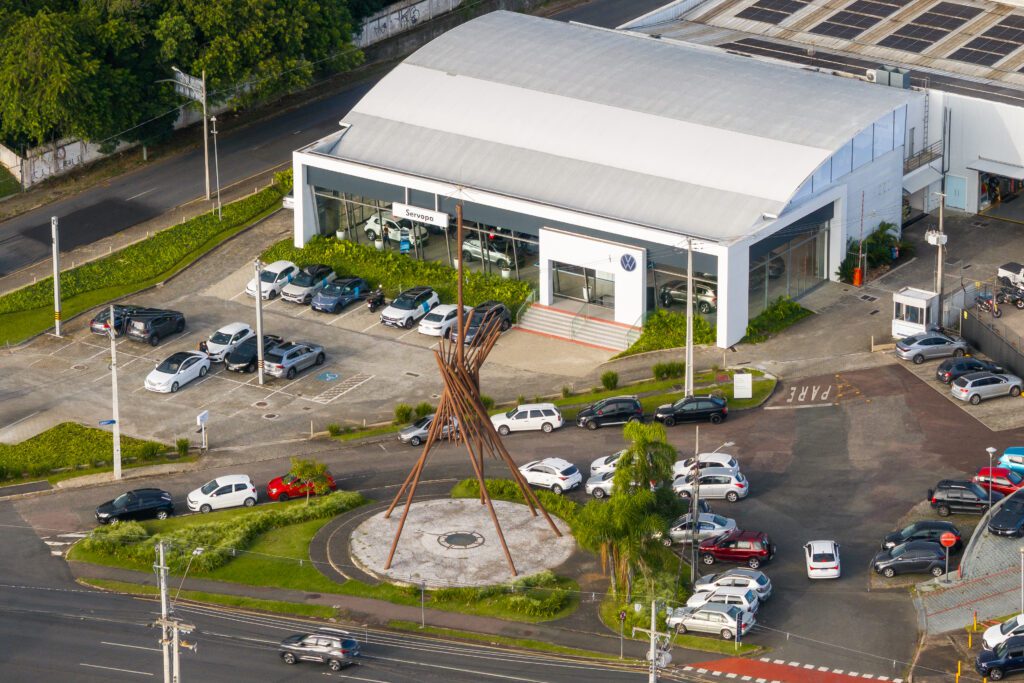 Vista aérea de uma moderna concessionária de automóveis com fachada branca e janelas de vidro, cercada por carros estacionados. Na frente, uma área circular apresenta uma escultura de metal alta. Estradas, vegetação e algumas pessoas podem ser vistas ao redor da concessionária. - Luiz Volpato Arquitetura
