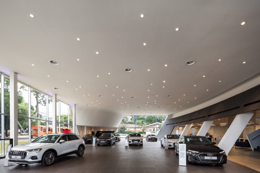 Showroom de carros espaçoso e moderno com teto alto e iluminação brilhante. Vários carros Audi, incluindo SUVs e sedãs, são exibidos em um piso de ladrilho brilhante. Grandes janelas à esquerda permitem a entrada de luz natural, e há um vislumbre de vegetação e uma rua do lado de fora. - Luiz Volpato Arquitetura