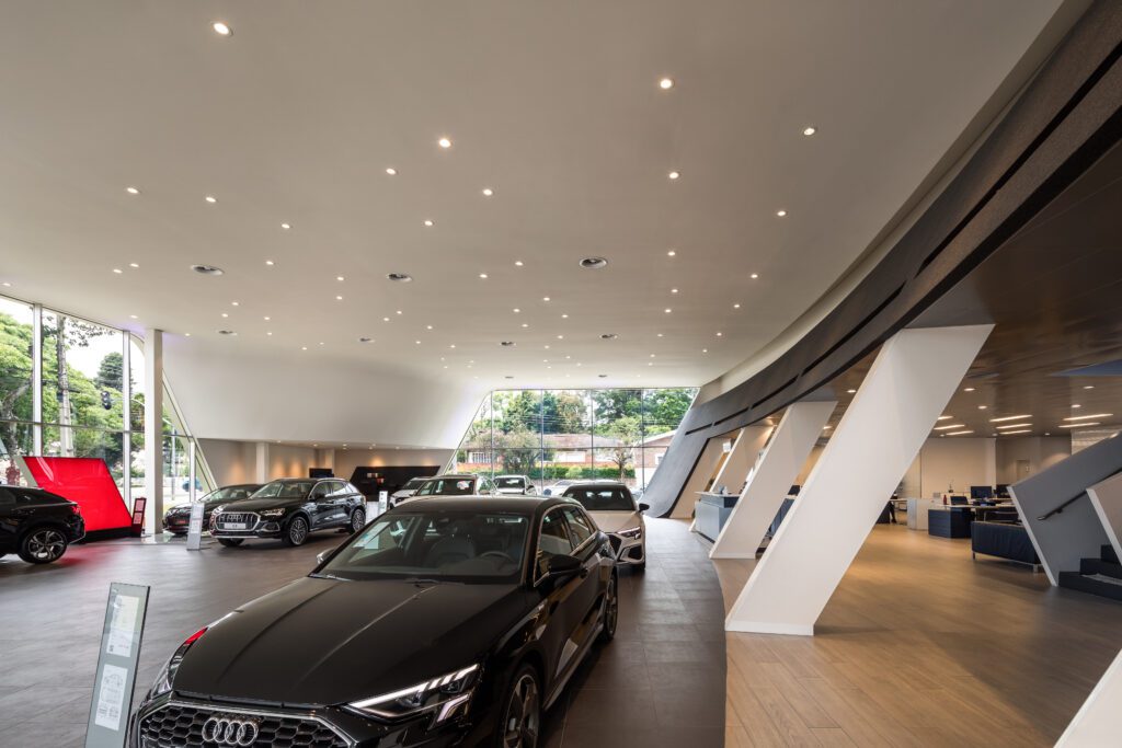 Interior de showroom de carros com um design elegante e moderno. Vários carros são exibidos, incluindo um Audi preto em primeiro plano. O espaço apresenta um teto curvo com iluminação embutida, grandes janelas revelando vegetação do lado de fora e colunas arquitetônicas ousadas ao longo de um lado. - Luiz Volpato Arquitetura