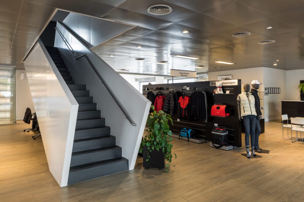 Showroom moderno com uma escada elegante à esquerda, levando para cima. Os displays apresentam roupas e acessórios, incluindo jaquetas e bonés, em prateleiras e manequins. Uma planta em vaso acrescenta vegetação. O espaço tem piso de madeira, iluminação brilhante e um design contemporâneo. - Luiz Volpato Arquitetura