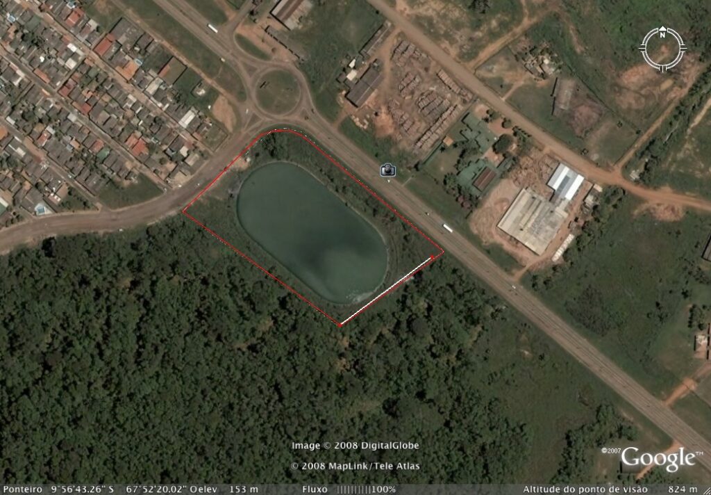 Vista aérea de um lago oval cercado por vegetação, cercado por uma estrada e prédios em dois lados. O lago é cercado por um contorno vermelho. À esquerda, há uma área residencial densamente povoada, e à direita, há estruturas industriais. - Luiz Volpato Arquitetura