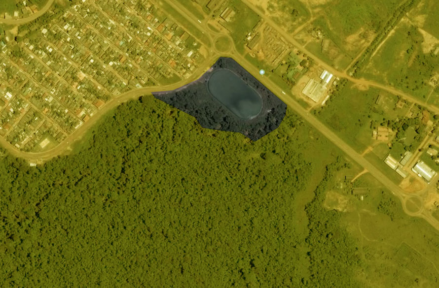 Vista aérea mostrando uma área florestal no canto inferior esquerdo, um pequeno lago perto do centro e casas residenciais no canto superior esquerdo. Estradas com prédios próximos margeiam o lago no canto superior direito. A paisagem é intercalada com vegetação e um padrão de ruas em forma de grade. - Luiz Volpato Arquitetura