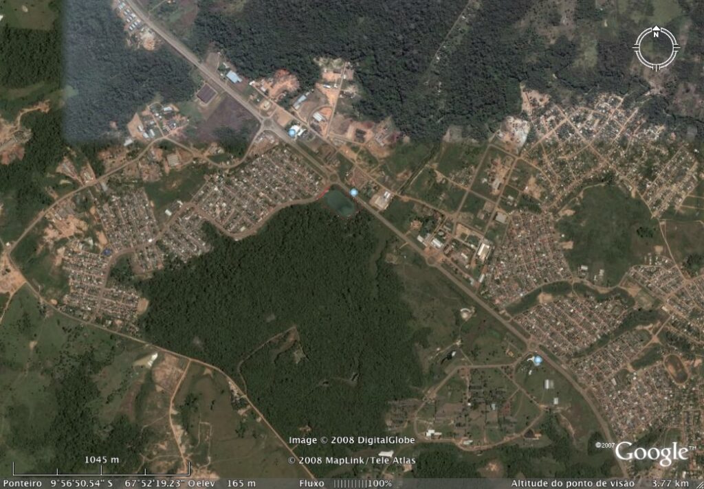 Uma imagem de satélite mostra uma cidade cercada por uma densa floresta verde. Estradas cruzam a área, com aglomerados de prédios e campos abertos espalhados por toda parte. O denso layout urbano da cidade contrasta com a paisagem natural ao redor. A imagem inclui detalhes de dados do mapa na parte inferior. - Luiz Volpato Arquitetura