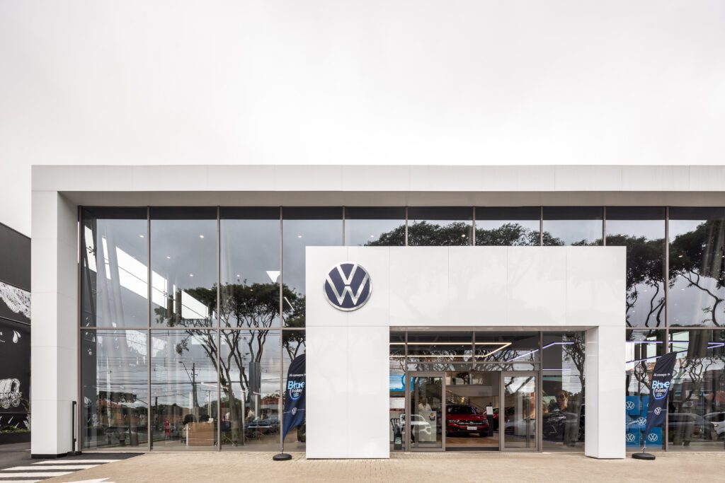 Exterior de uma concessionária de carros moderna com uma grande fachada de vidro e um logotipo proeminente da Volkswagen perto da entrada. No interior, os carros são visíveis com faixas promocionais azuis. O céu está nublado, e as árvores são refletidas nas janelas, adicionando ao visual contemporâneo. - Luiz Volpato Arquitetura