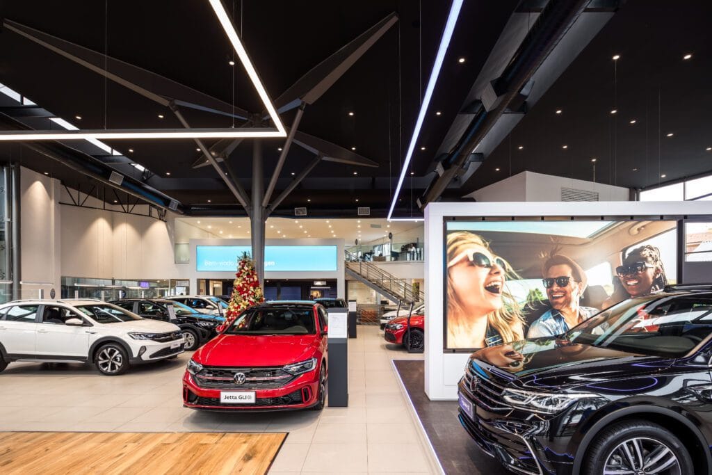 Um showroom de carros moderno com vários modelos, incluindo um Jetta GLI vermelho em destaque. Outros carros são brancos e pretos. Grandes telas digitais mostram imagens de pessoas alegres em um carro. O espaço é bem iluminado com elementos de design elegantes e contemporâneos e uma árvore de Natal visível. - Luiz Volpato Arquitetura