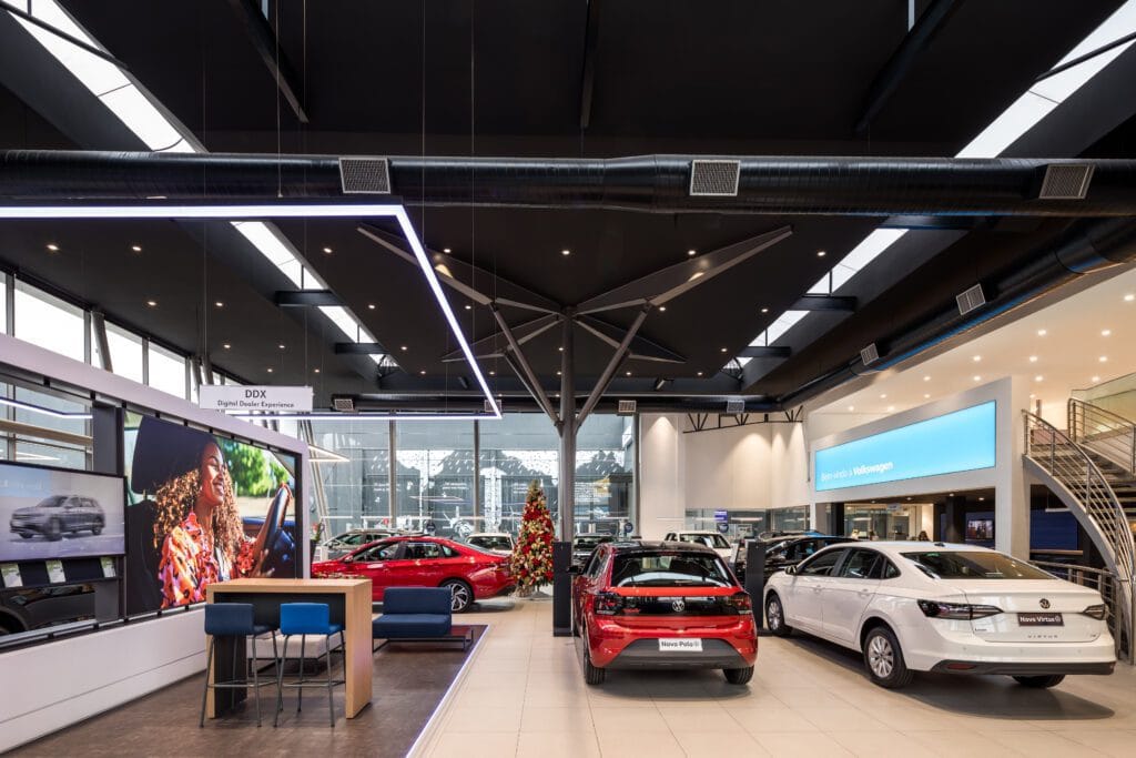 Um showroom de carros moderno com vários carros em exposição. O espaço tem um teto alto com claraboias e iluminação de trilho. Uma tela digital mostra uma mulher dirigindo. Há uma área de estar com bancos e uma árvore de Natal ao fundo. Escadas levam a um nível superior. - Luiz Volpato Arquitetura