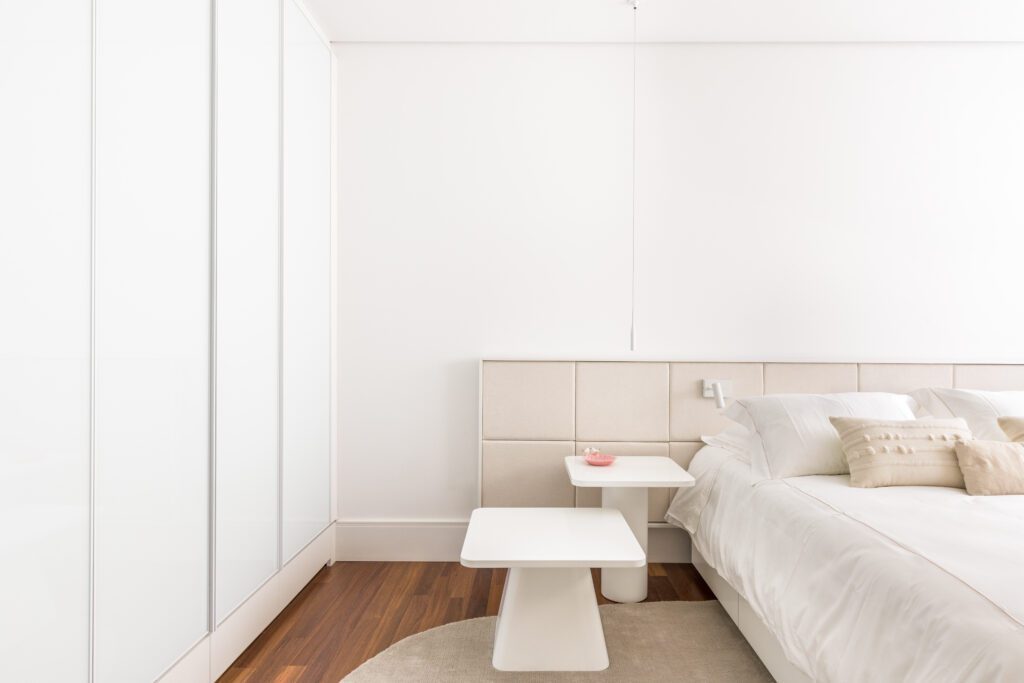 Um quarto luminoso e minimalista apresenta uma cama com painéis de cabeceira acolchoados em bege e roupa de cama branca. Duas mesinhas laterais brancas, do tipo pedestal, estão sobre um tapete circular no piso de madeira escura, ao lado de uma parede com armários brancos que vão do chão ao teto. - Luiz Volpato Arquitetura