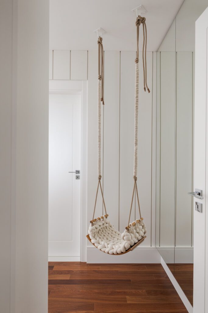 Uma cadeira suspensa decorativa feita de corda trançada grossa, na cor creme, pendurada no teto. Ela fica em um corredor minimalista com paredes brancas com painéis verticais, piso de madeira escura e um espelho que vai do chão ao teto. - Luiz Volpato Arquitetura