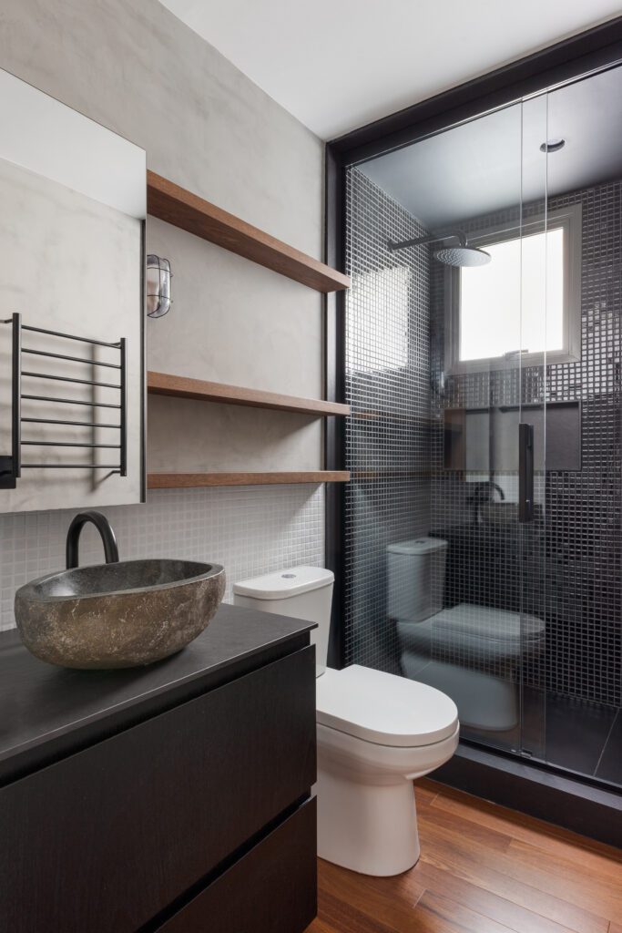 Uma pia de pedra repousa sobre um lavatório escuro ao lado de um vaso sanitário branco. Atrás deles, um box com divisórias de vidro apresenta paredes revestidas com azulejos de mosaico escuros, e um piso de madeira se estende por todo o cômodo. Duas prateleiras suspensas de madeira estão fixadas em uma parede cinza-clara. - Luiz Volpato Arquitetura