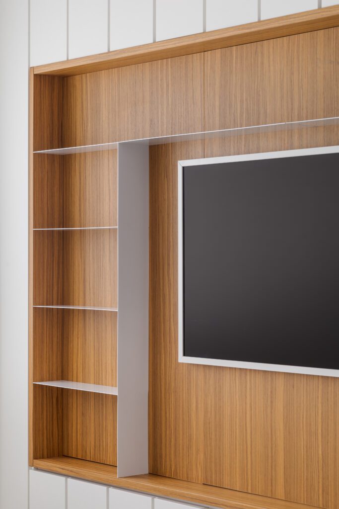 Uma televisão fixada na parede está posicionada no centro de uma parede de destaque revestida com painéis de madeira. À sua esquerda, estantes verticais brancas e finas oferecem espaço de armazenamento embutido. O design clean e moderno cria um contraste entre o tom marrom aconchegante do veio da madeira e as bordas brancas e bem definidas das prateleiras. - Luiz Volpato Arquitetura