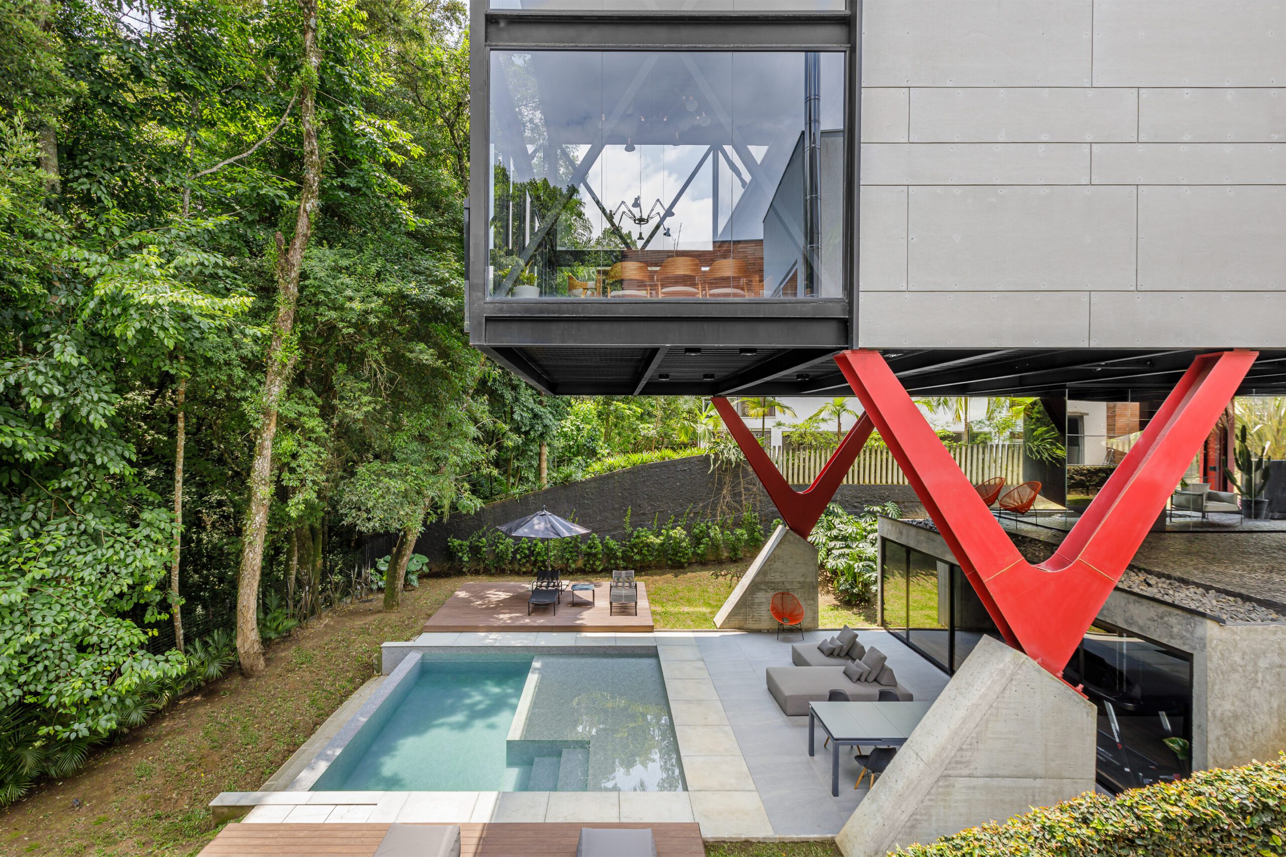 Casa moderna cercada por vegetação exuberante. A seção elevada apresenta grandes janelas de vidro e suportes vermelhos em forma de V. Abaixo, há um pátio com móveis de exterior, uma piscina e uma área para banhos de sol com espreguiçadeiras e um guarda-sol. O cenário é uma floresta densa, criando uma atmosfera serena. - Luiz Volpato Arquitetura