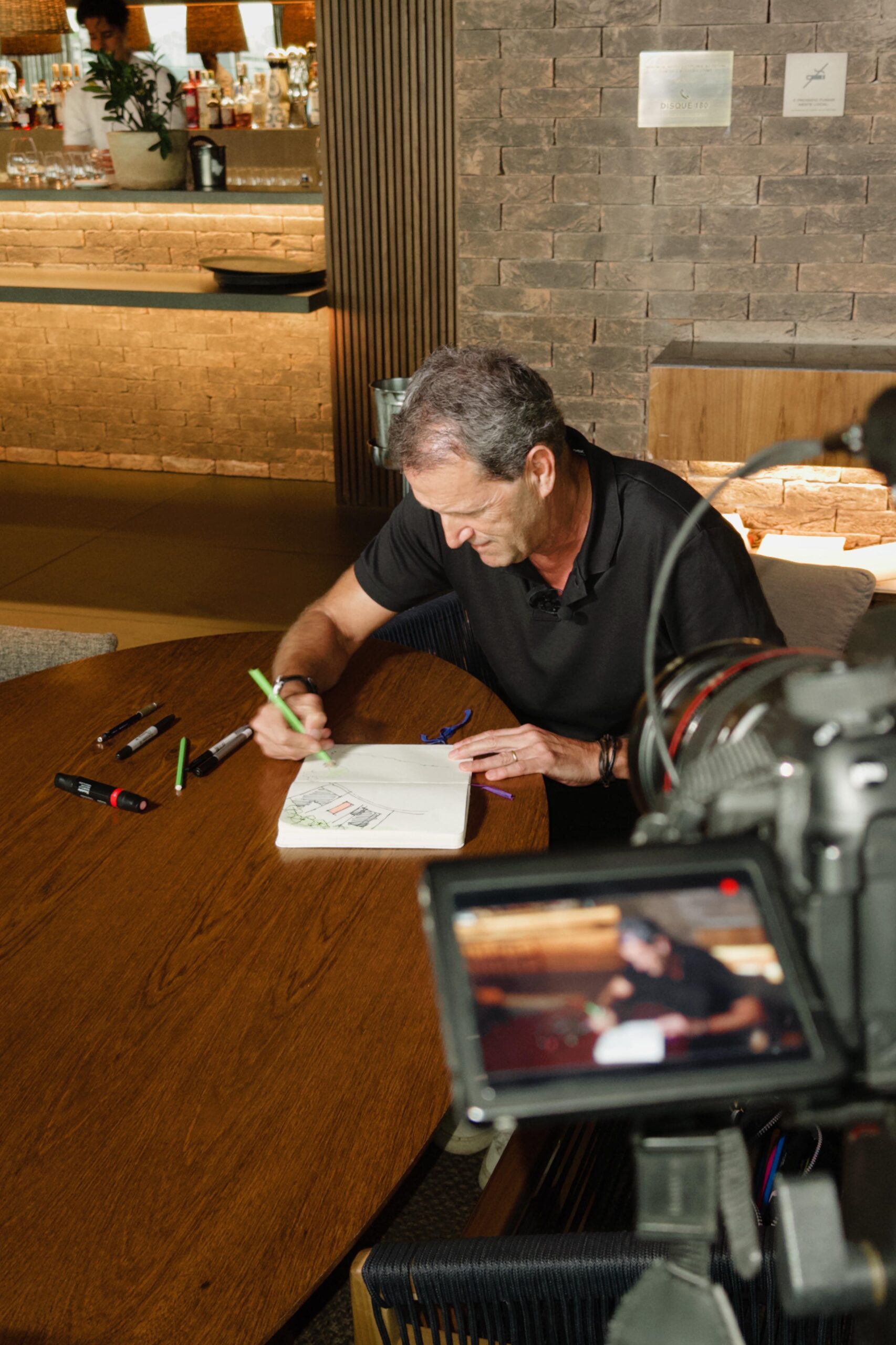 Um homem de camisa preta está sentado em uma mesa de madeira, desenhando em um caderno com marcadores coloridos. Uma câmera de filmagem está posicionada em primeiro plano, capturando a cena. O fundo apresenta uma parede de tijolos e uma pessoa desfocada em branco perto de uma área de bar. - Luiz Volpato Arquitetura