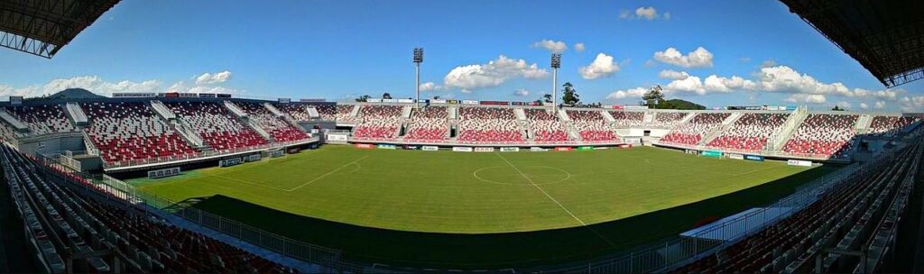 Vista panorâmica de um estádio de futebol vazio com grama verde, cercado por arquibancadas com assentos vermelhos e brancos. Holofotes se elevam acima, e um céu azul claro com nuvens dispersas é visível. O estádio é bem iluminado, e as áreas de assentos formam padrões com as cores. - Luiz Volpato Arquitetura