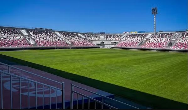 Um estádio de futebol com uma área de assentos vazia, com assentos pretos, brancos e vermelhos. O campo verde bem cuidado fica no centro sob um céu azul claro. O estádio é cercado por holofotes altos e tem uma grande pista ao redor do campo. - Luiz Volpato Arquitetura