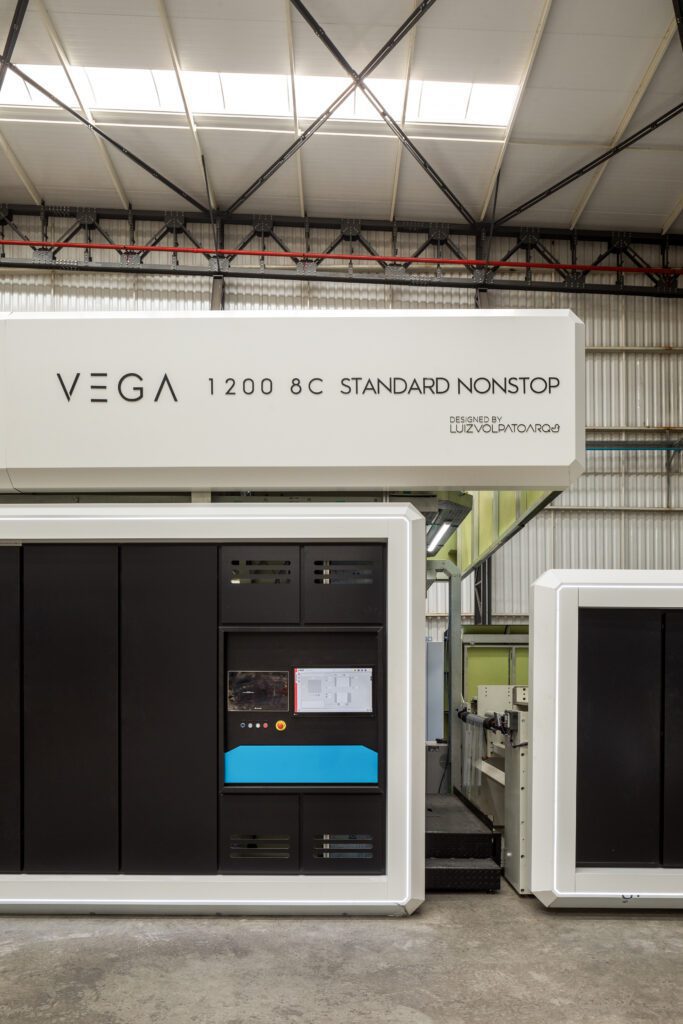 Uma grande máquina de impressão industrial, rotulada "Vega 1200 8C Standard Nonstop", está instalada dentro de um armazém espaçoso com paredes de metal corrugado e teto alto. A máquina possui painéis pretos, um display digital com controles e parece estar em um ambiente limpo e organizado. - Luiz Volpato Arquitetura