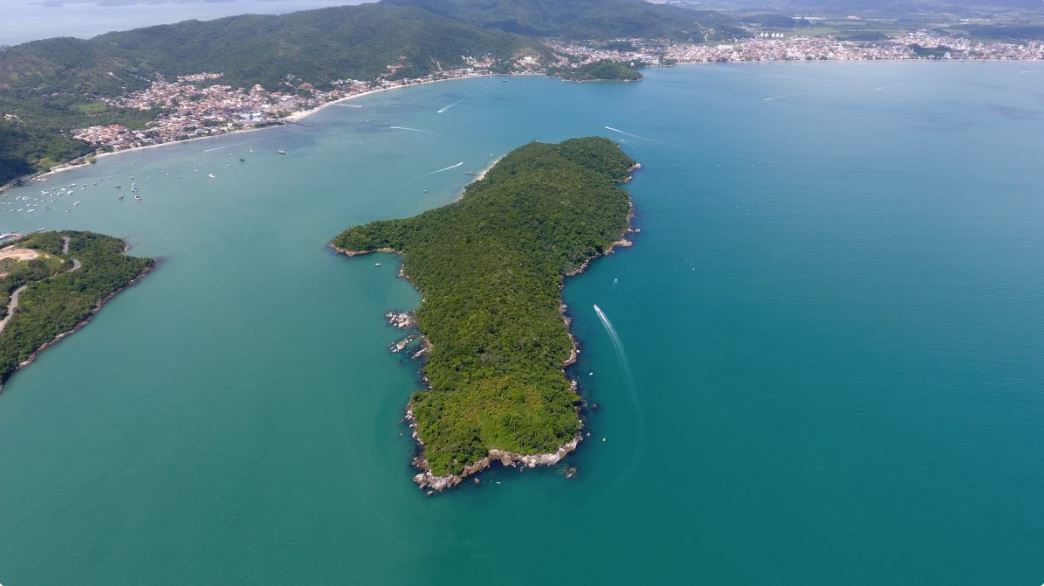 Vista aérea de uma península verdejante e exuberante que se estende até um mar calmo e turquesa. O litoral curva-se suavemente com barcos espalhados perto da costa. A terra é densamente florestada, e uma cidade distante com prédios e estradas é visível perto da costa, emoldurada por colinas ondulantes. - Luiz Volpato Arquitetura