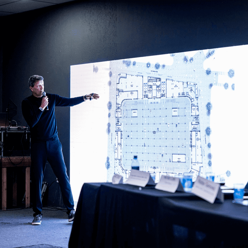 Um homem segurando um microfone está de pé e gesticula em direção a um grande visor digital que mostra uma planta arquitetônica. Em primeiro plano, uma mesa com garrafas de água e placas de identificação é visível, sugerindo uma apresentação ou reunião profissional. - Luiz Volpato Arquitetura