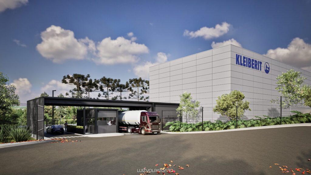 Um prédio industrial moderno com o logotipo “Kleiberit”, cercado por árvores. Um caminhão e um carro estão estacionados em uma entrada cercada, com uma guarita. O céu está quase totalmente limpo, com algumas nuvens dispersas. Há algumas folhas espalhadas pela estrada. - Luiz Volpato Arquitetura