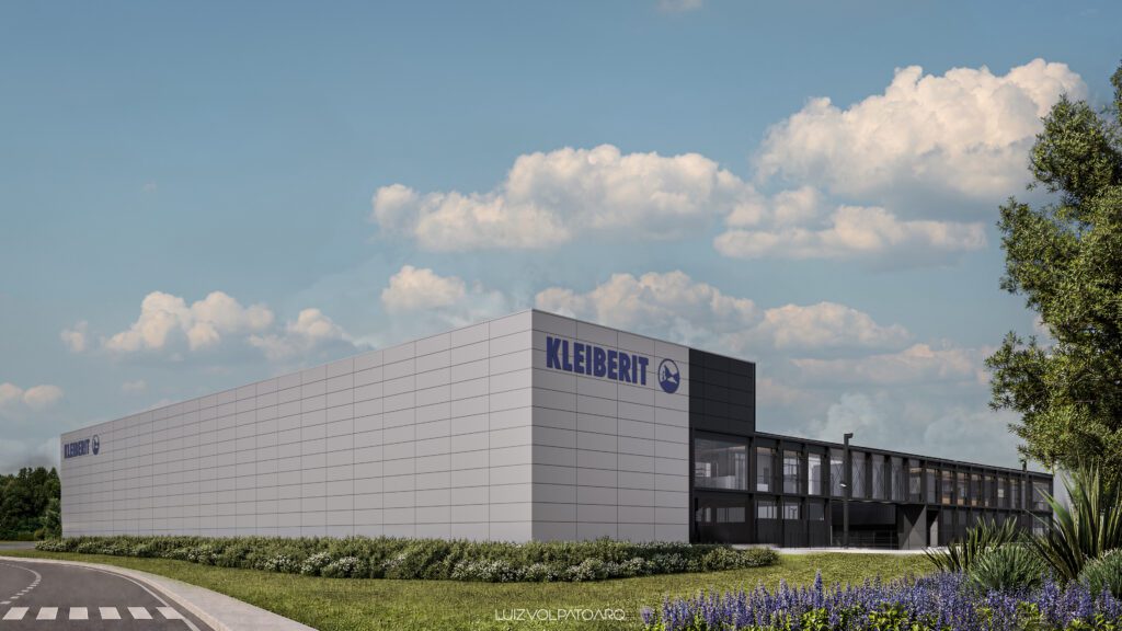 Um grande e moderno prédio industrial cinza com a inscrição “KLEIBERIT”, cercado por gramados bem aparados, arbustos e flores roxas, sob um céu azul com nuvens esparsas. Uma rua com uma faixa de pedestres passa pelo primeiro plano. - Luiz Volpato Arquitetura