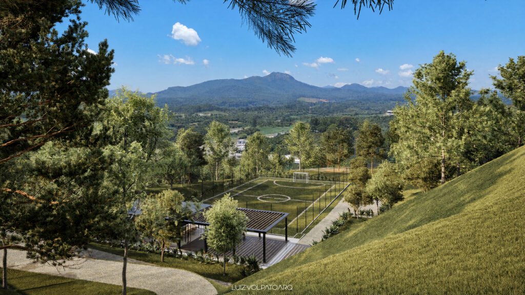 Um campo de futebol cercado fica em meio a árvores verdes e colinas onduladas. Uma cordilheira é visível ao fundo, sob um céu azul com nuvens esparsas. Em primeiro plano, há uma pequena área de descanso com sombra e um caminho sinuoso. - Luiz Volpato Arquitetura