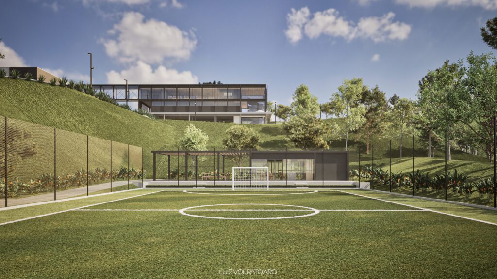 Um moderno campo de futebol ao ar livre, com linhas brancas, estende-se sobre a grama verde, cercado por uma cerca de rede. Ao fundo, há dois edifícios elegantes com paredes de vidro em uma colina, rodeados por árvores frondosas, arbustos e um céu azul com nuvens esparsas. - Luiz Volpato Arquitetura
