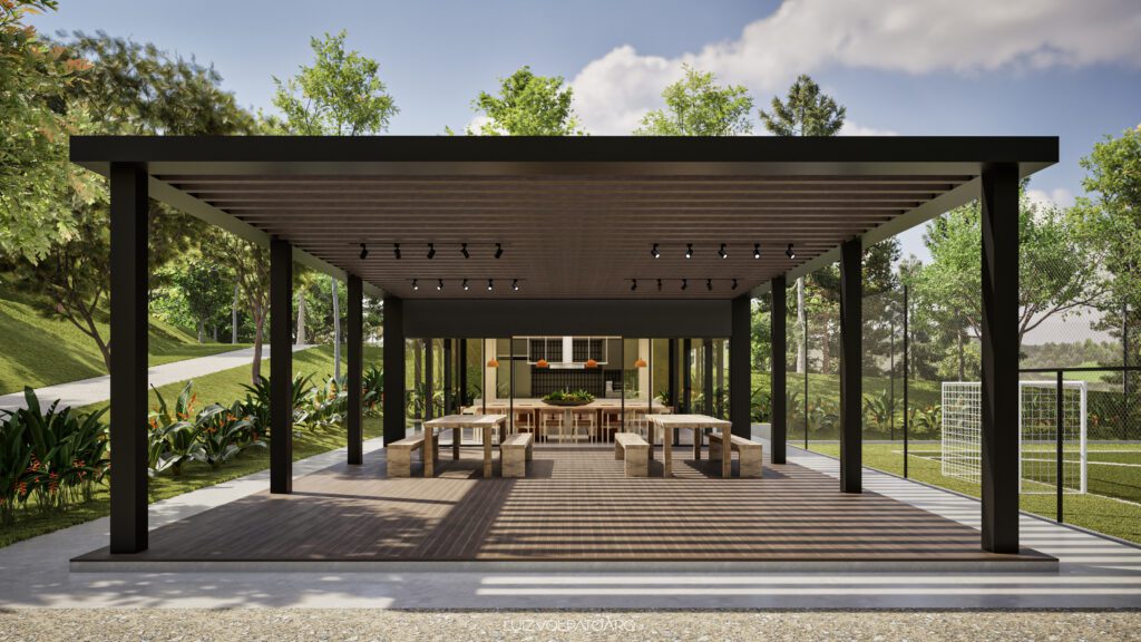 Um pavilhão moderno ao ar livre com telhado em pérgula preta, piso de madeira, longas mesas de jantar, bancos, luzes decorativas e vegetação. Uma trave de futebol é visível no gramado à direita. Árvores frondosas e o céu azul compõem o fundo. - Luiz Volpato Arquitetura