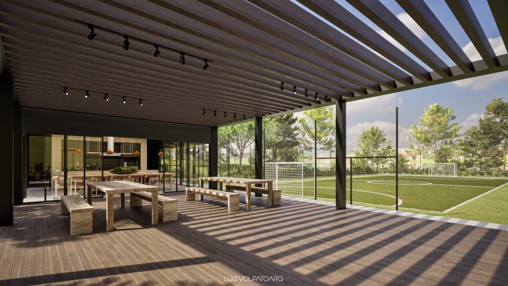 Um pátio externo coberto, com bancos e mesas de madeira, fica de frente para um campo de futebol cercado, sob um céu limpo. A luz do sol cria sombras listradas através do telhado de ripas. Árvores e arbustos de um verde exuberante cercam esse espaço recreativo moderno e aberto. - Luiz Volpato Arquitetura