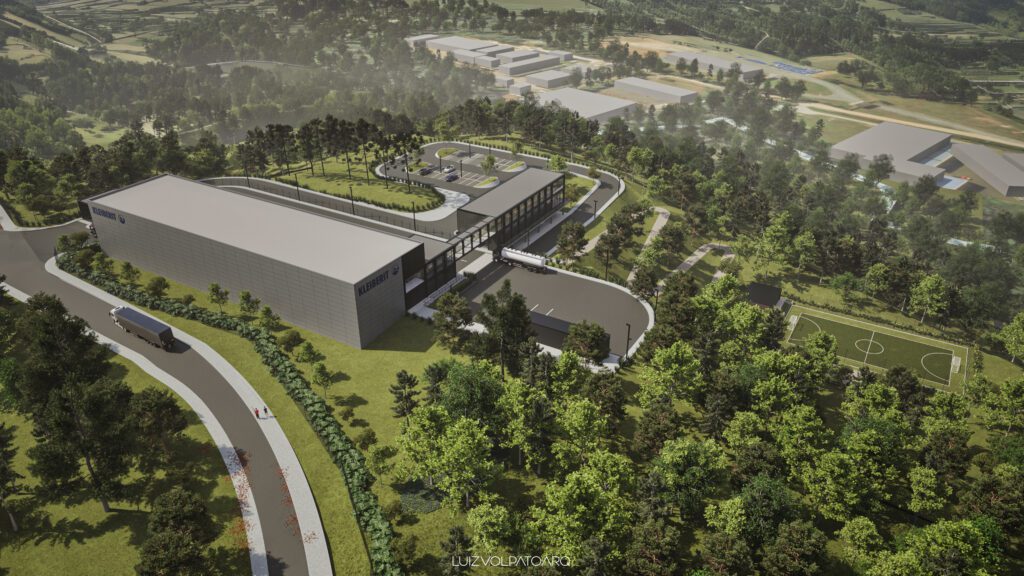 Um grande e moderno prédio industrial cinza, com um telhado verde e uma entrada sinuosa, está situado em meio a árvores densas. Caminhões estão estacionados nas proximidades. À direita, há um pequeno campo de futebol, e vários prédios são visíveis no fundo, em meio à floresta. - Luiz Volpato Arquitetura