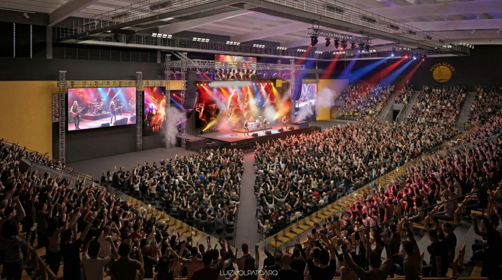 Uma arena coberta lotada recebe um show animado. Luzes coloridas iluminam o palco, onde uma banda se apresenta. O público está de pé, aplaudindo com os braços erguidos. Telões grandes ficam de cada lado do palco, e o local possui tetos altos e arquibancadas em degraus. - Luiz Volpato Arquitetura