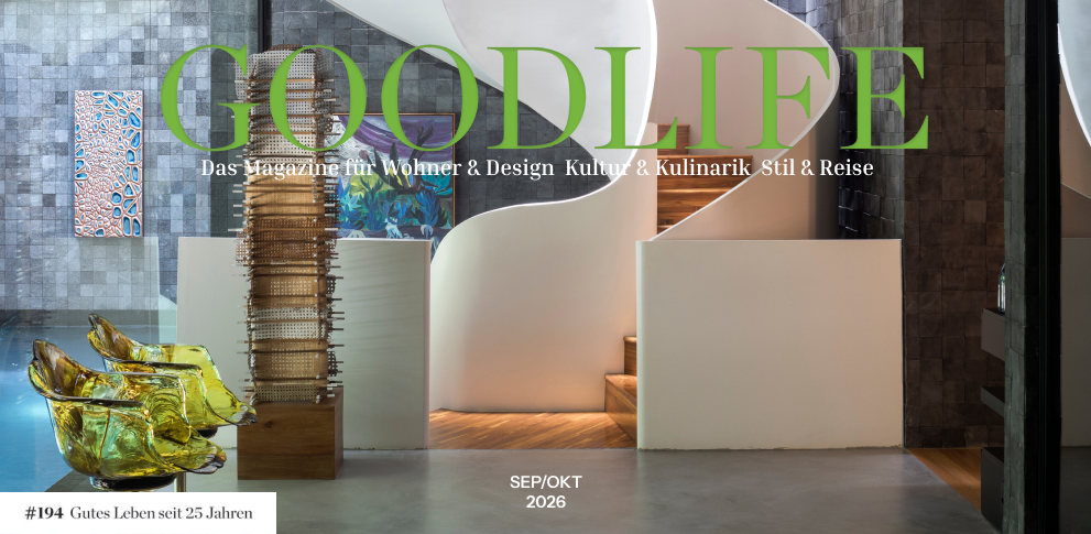 Interior moderno com uma escada branca em espiral, obras de arte abstratas nas paredes de pedra e cadeiras amarelas translúcidas. A palavra “GOODLIFE” aparece em letras verdes grandes. Aparecem o subtítulo e a data “SET/OUT 2026”, com uma etiqueta na parte inferior esquerda comemorando os 25 anos de publicação. - Luiz Volpato Arquitetura