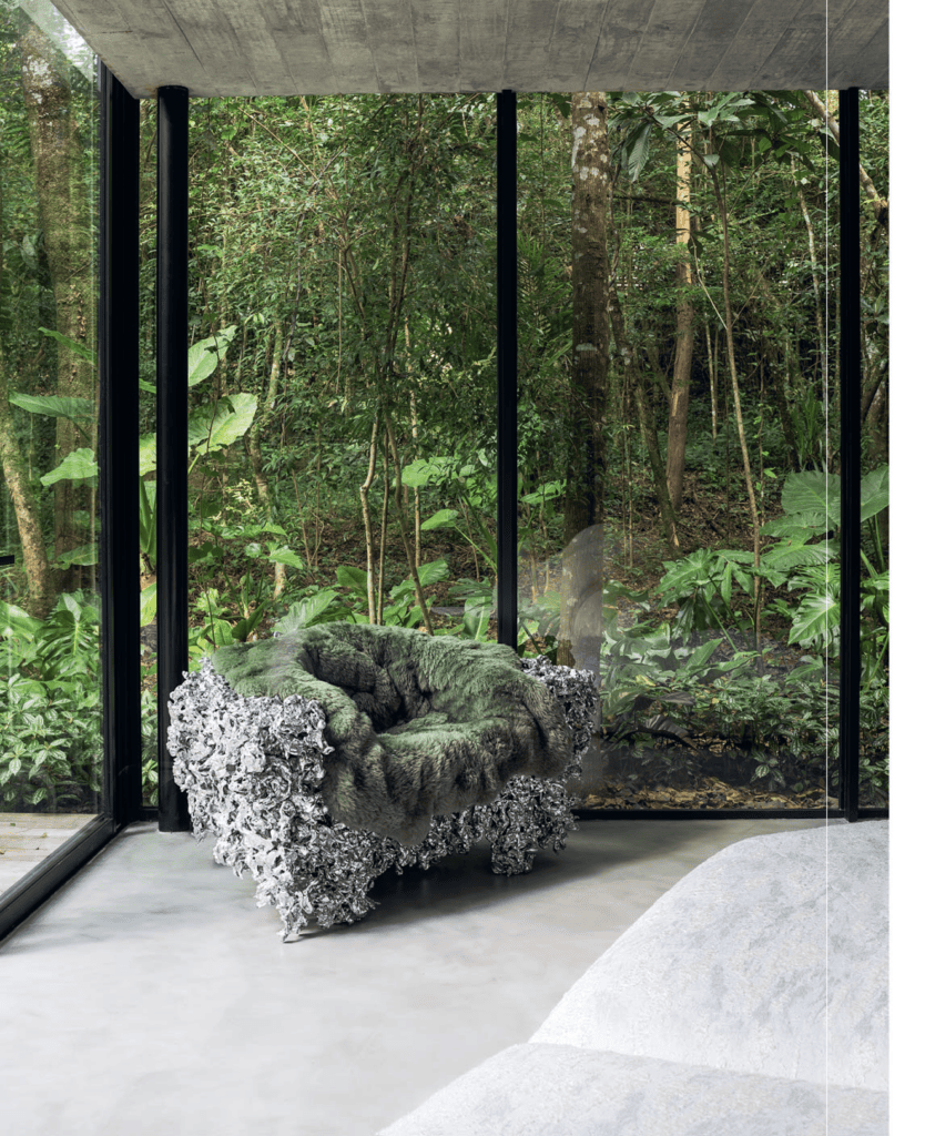 Uma cadeira escultural com base metálica e tecido macio na cor verde-musgo está posicionada em uma sala moderna com paredes de vidro. Uma exuberante floresta verde preenche a vista lá fora. O teto de concreto e o piso cinza liso criam uma atmosfera elegante e minimalista. - Luiz Volpato Arquitetura