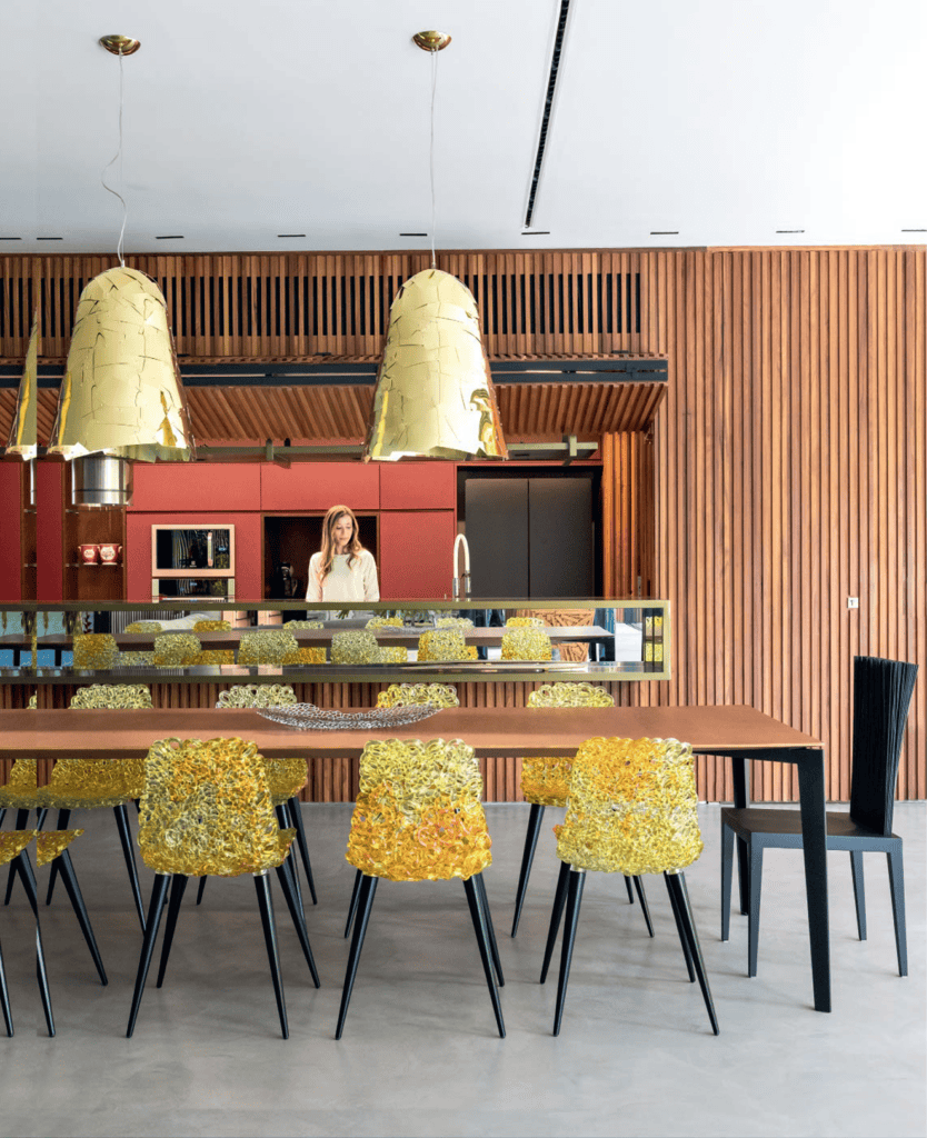 Uma sala de jantar moderna com paredes revestidas de painéis de madeira, uma longa mesa de madeira e cadeiras exclusivas de cor amarela com textura. Três grandes luminárias pendentes douradas estão suspensas no teto. Uma mulher está em pé perto de um revestimento vermelho na área da cozinha aberta, ao fundo. - Luiz Volpato Arquitetura