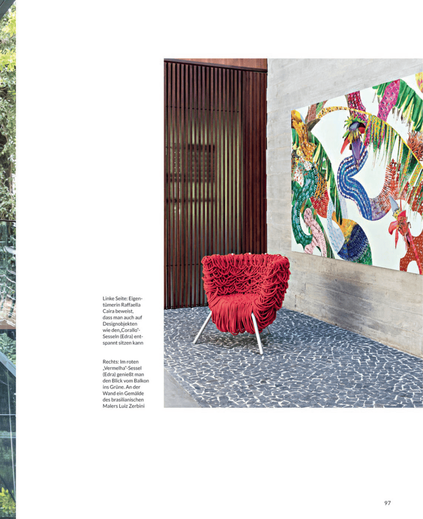 Um interior moderno com uma cadeira vermelha com babados, uma parede cinza texturizada, uma grande obra de arte colorida retratando animais abstratos e uma divisória de ripas de madeira. O piso apresenta um padrão de mosaico de pedra, e é possível avistar vegetação através de uma janela de vidro à esquerda. - Luiz Volpato Arquitetura