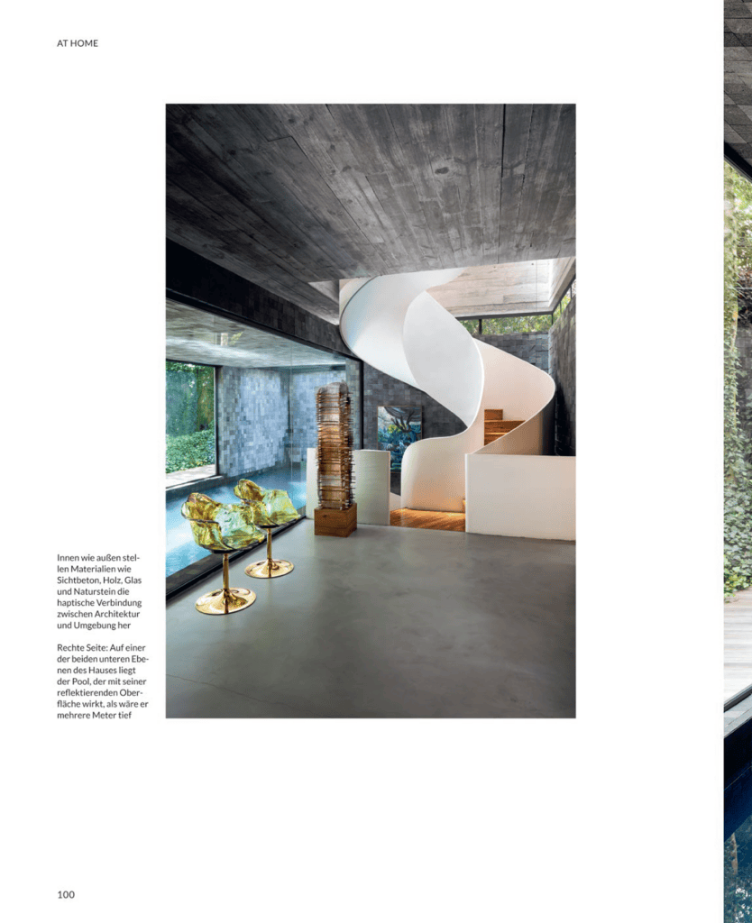 Um interior moderno com paredes e teto de concreto, uma escada em espiral branca, grandes janelas de vidro, esculturas douradas sobre pedestais e uma piscina visível em um dos lados. Do lado de fora, vê-se uma vegetação exuberante, que contrasta com o design minimalista e industrial. - Luiz Volpato Arquitetura
