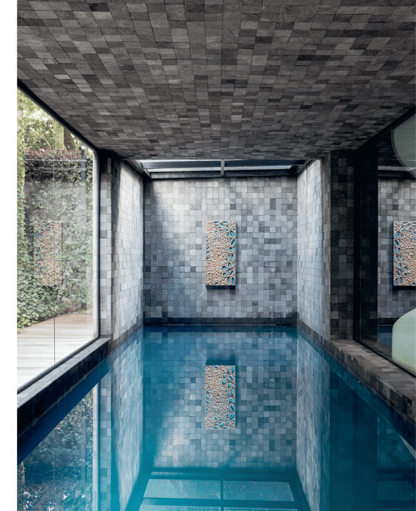 Piscina coberta com azulejos de pedra cinza nas paredes e no teto, refletindo-se na água azul cristalina. Uma grande janela de vidro mostra a vegetação do lado de fora. Duas obras de arte abstratas coloridas estão penduradas nas paredes traseira e lateral. O espaço transmite uma sensação de modernidade e tranquilidade. - Luiz Volpato Arquitetura