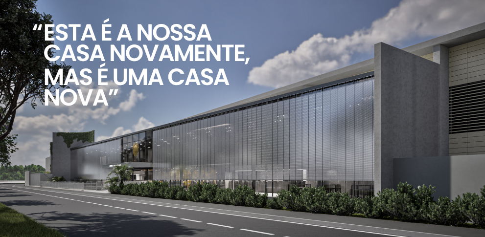 Um edifício moderno com grandes painéis de vidro, vegetação na base e uma rua em frente. O céu está parcialmente nublado. À esquerda, uma inscrição em branco em português diz: “ESTA É A NOSSA CASA NOVAMENTE, MAS É UMA CASA NOVA.” Este exemplo marcante de #arquiteturaesportiva reflete o design inovador da #luizvolpatoarquitetura, combinando elegância e funcionalidade na paisagem urbana de Uberlândia. - Luiz Volpato Arquitetura